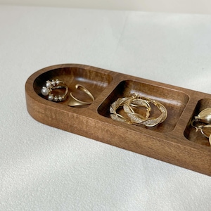 May include: A wooden jewelry tray with three compartments, holding various earrings. The earrings include a pair with pearls, a gold ear cuff, and a pair of gold hoop earrings with clear stones. The tray is made of dark brown wood.