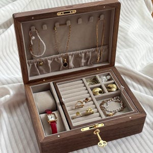 Puede incluir: Un joyero rectangular de madera con acabado marr&oacute;n oscuro e interior de terciopelo beige. La tapa tiene ganchos para collares, mientras que la base contiene un coj&iacute;n para reloj, ranuras para anillos y compartimentos con anillos de oro y pulseras.