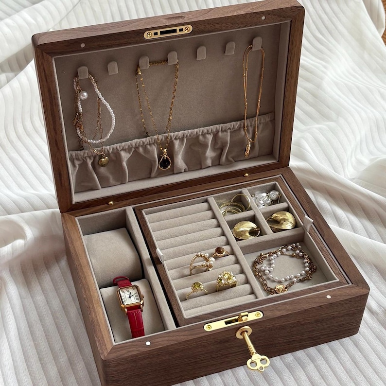 Puede incluir: Un joyero rectangular de madera con acabado marr&oacute;n oscuro e interior de terciopelo beige. La tapa tiene ganchos para collares, mientras que la base contiene un coj&iacute;n para reloj, ranuras para anillos y compartimentos con anillos de oro y pulseras.