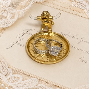 May include: A gold-toned metal ring dish featuring a small dog figurine holding a clear barbell. The circular tray has a textured, hammered surface and holds two silver-colored rings, one with a large central stone and one with channel-set stones.