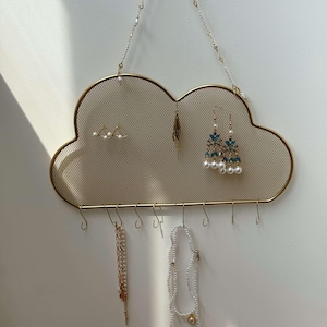Op de afbeelding: Een wolk-vormige, goudkleurige metalen sieraden organizer met een gaas achterkant. De organizer heeft haken aan de onderkant om kettingen en oorbellen op te hangen. Verschillende sieraden, waaronder oorbellen en kettingen, worden getoond.