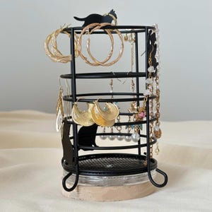 Peut inclure: Organisateur de bijoux en métal noir avec un motif de silhouette de chat. L'organisateur contient diverses boucles d'oreilles dorées, notamment des créoles et des modèles pendants. La base est un bocal transparent avec une base en bois brun clair.