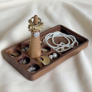 May include: A rectangular wooden jewelry organizer tray featuring a central conical ring holder and multiple circular recessed compartments. The tray holds various rings on the cone, a white pearl necklace, and assorted earrings and small jewelry pieces.