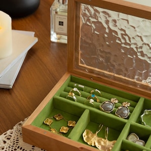 May include: A wooden jewellery box with a glass lid, displaying rings, earrings, and a bracelet. The interior is lined with green velvet, contrasting with the gold and silver jewellery. The box is open, revealing organised compartments for storage.