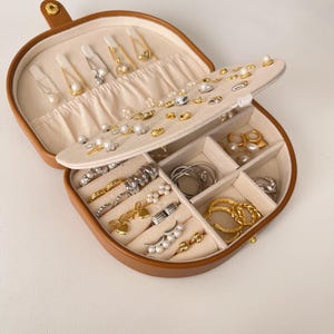 Pode incluir: Uma caixa de joias de couro marrom, aberta para exibir vários brincos, anéis e outras peças de joalheria. A caixa tem um interior bege com vários compartimentos e ranhuras. A joalheria inclui tons dourados e prateados, com pérolas e outros elementos decorativos.