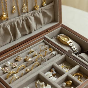 Puede incluir: Un joyero de madera con interior de terciopelo beige que contiene varios collares dorados colgados de la tapa, junto con m&uacute;ltiples anillos, pendientes y un reloj de pulsera dorado con correa de eslabones met&aacute;licos en los compartimentos.