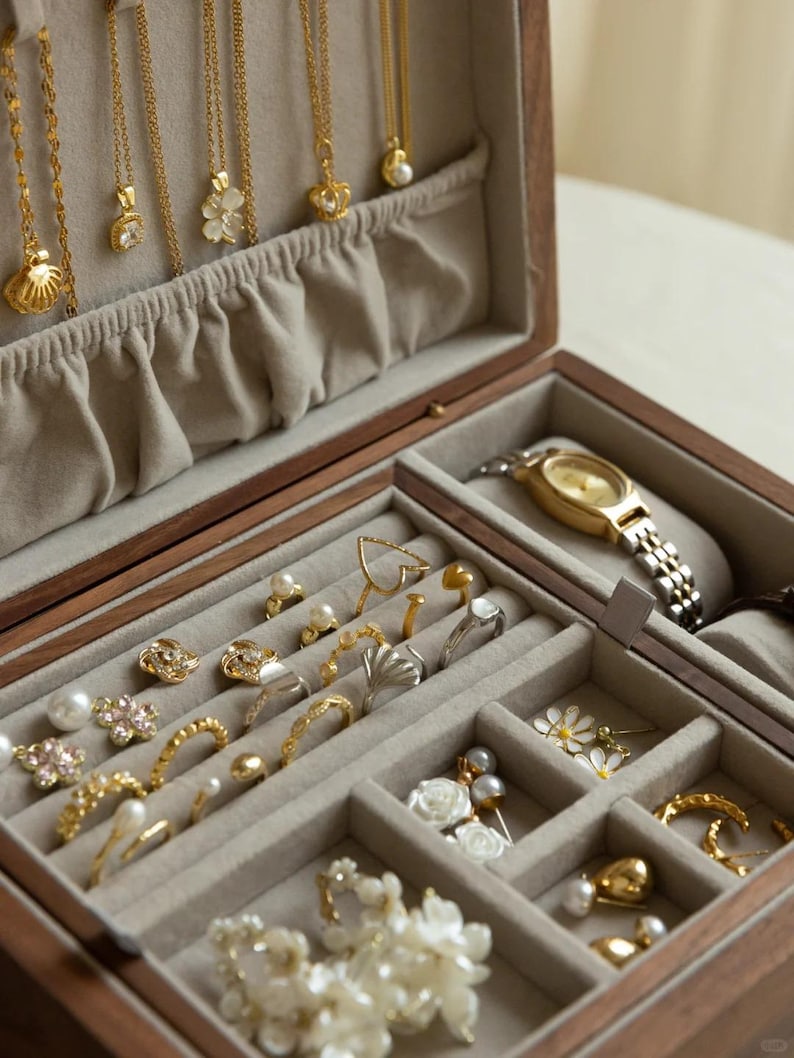Puede incluir: Un joyero de madera con interior de terciopelo beige que contiene varios collares dorados colgados de la tapa, junto con m&uacute;ltiples anillos, pendientes y un reloj de pulsera dorado con correa de eslabones met&aacute;licos en los compartimentos.