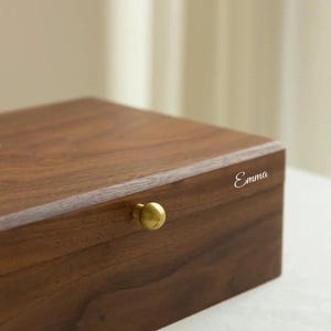 Puede incluir: Una caja rectangular de madera marr&oacute;n oscuro con acabado liso, que presenta un peque&ntilde;o pomo redondo de metal dorado en el panel frontal. El nombre Emma est&aacute; grabado en una fuente cursiva blanca en la esquina superior derecha del borde frontal.
