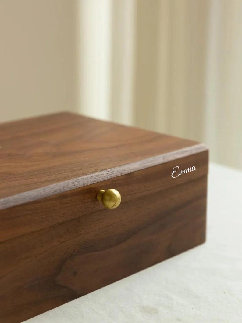 Puede incluir: Una caja rectangular de madera marr&oacute;n oscuro con acabado liso, que presenta un peque&ntilde;o pomo redondo de metal dorado en el panel frontal. El nombre Emma est&aacute; grabado en una fuente cursiva blanca en la esquina superior derecha del borde frontal.