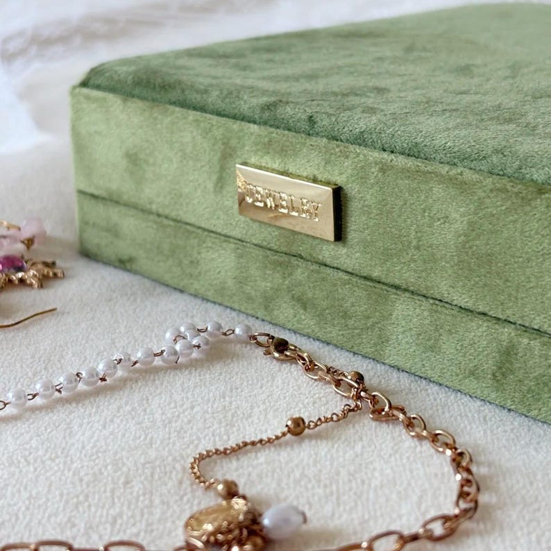 Może przedstawiać: Szałwiowa zielona aksamitna szkatułka na biżuterię ze złotą prostokątną tabliczką znamionową z napisem "JEWELRY". Złoty naszyjnik z perłową zawieszką i biały naszyjnik z pereł są na pierwszym planie.
