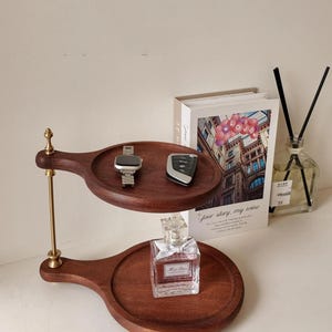 May include: A two-tiered wooden serving tray with a gold-colored support. The top tray holds a silver watch and a car key fob. The bottom tray displays a perfume bottle. A book and a diffuser are in the background.