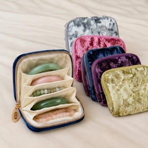 May include: A collection of small, square, zippered velvet jewelry cases in gray, pink, navy, purple, and olive green. One navy case is open, revealing cream-colored fabric dividers holding jade-colored bangles and beaded bracelets.