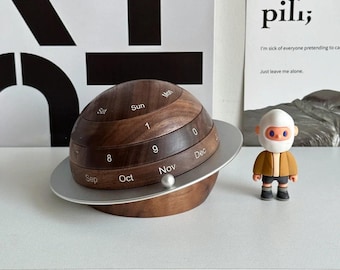 May include: A wooden perpetual calendar in the shape of a planet with a silver ring. The calendar displays months and numbers. A small astronaut figurine stands nearby. The background includes a poster with the word "pili;" and the text "I'm sick of everyone pretending to care. Just leave me alone."