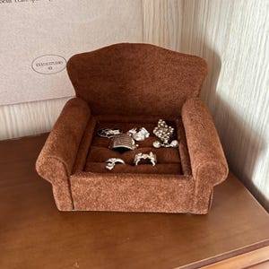 Puede incluir: Un pequeño expositor de anillos en forma de sofá de terciopelo marrón, que muestra varios anillos. El expositor tiene respaldo curvo y reposabrazos. Los anillos son plateados y presentan diversos diseños, incluyendo patrones florales y geométricos.
