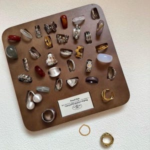 Puede incluir: Una colección de anillos exhibidos en una tabla de madera marrón. Los anillos varían en estilo, incluyendo bandas, engastes de piedras preciosas y diseños decorativos. Algunos anillos presentan perlas, mariposas y motivos florales.