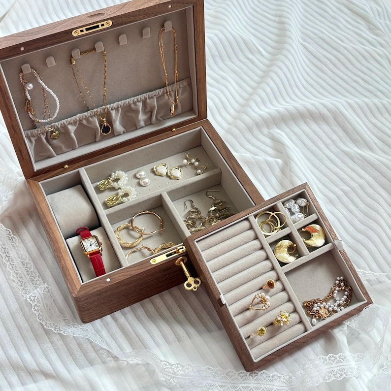 Puede incluir: Un joyero rectangular de madera con acabado en nogal oscuro e interior de terciopelo beige. La tapa tiene ganchos para collares, mientras que la base contiene compartimentos para pendientes, anillos, un reloj con correa de cuero rojo y una llave dorada.