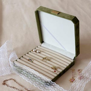 May include: Open green velvet jewellery box with a cream interior, displaying rings and earrings. The box is open, revealing a cream interior with slots for rings. A white lace ribbon and a gold necklace are on the side.