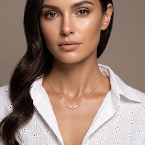 May include: A gold necklace with a pendant in a curved shape is worn over a white eyelet shirt. The model has dark hair and is looking at the camera. The background is a solid neutral color.