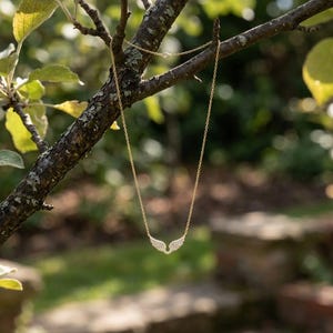 May include: A delicate gold necklace with a pendant shaped like angel wings, adorned with small, sparkling stones. The necklace hangs from a tree branch against a blurred background of green foliage and sunlight.