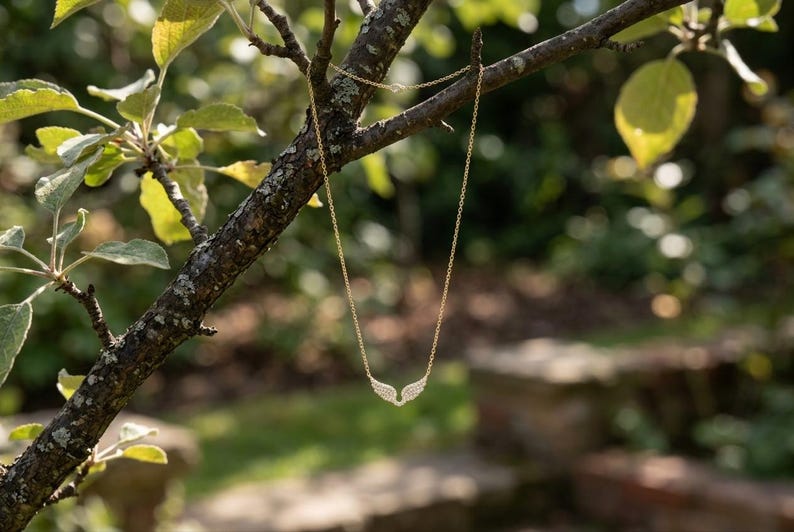 May include: A delicate gold necklace with a pendant shaped like angel wings, adorned with small, sparkling stones. The necklace hangs from a tree branch against a blurred background of green foliage and sunlight.