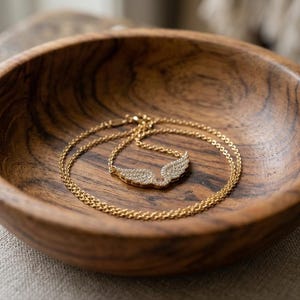 May include: A gold-colored necklace with a wing pendant, encrusted with small, sparkling stones, rests inside a wooden bowl. The bowl has a rich, dark wood grain. The necklace is delicate and the pendant is shaped like angel wings.