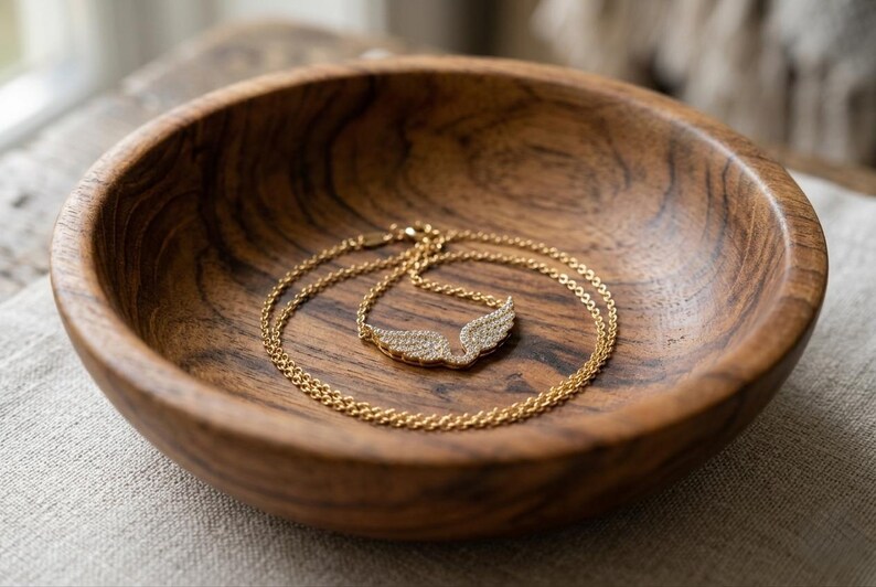 May include: A gold-colored necklace with a wing pendant, encrusted with small, sparkling stones, rests inside a wooden bowl. The bowl has a rich, dark wood grain. The necklace is delicate and the pendant is shaped like angel wings.