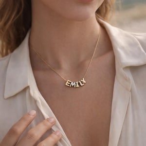 Könnte beinhalten: Eine goldfarbene Halskette mit dem Namen "EMILY" in Großbuchstaben. Die Halskette wird von einer Person getragen, die ein cremefarbenes Hemd mit Kragen trägt. Die Buchstaben sind einzeln und durch eine zarte Kette verbunden.