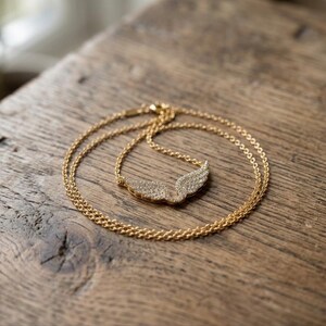 May include: A gold-colored necklace with a wing pendant, resting on a wooden surface. The pendant is encrusted with small, sparkling stones. The chain is delicate and the overall design is elegant.