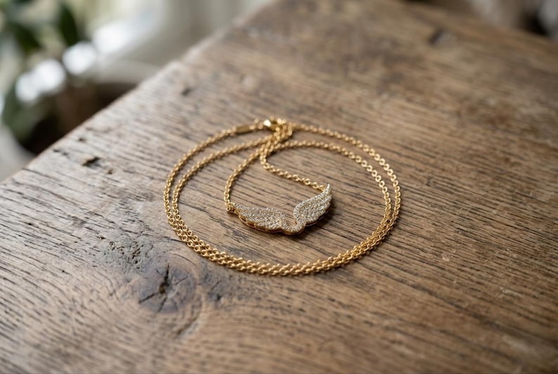 May include: A gold-colored necklace with a wing pendant, resting on a wooden surface. The pendant is encrusted with small, sparkling stones. The chain is delicate and the overall design is elegant.
