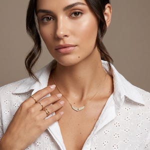 May include: A gold necklace with a wing-shaped pendant and several gold rings. The model is wearing a white eyelet shirt. The background is a solid, neutral color.