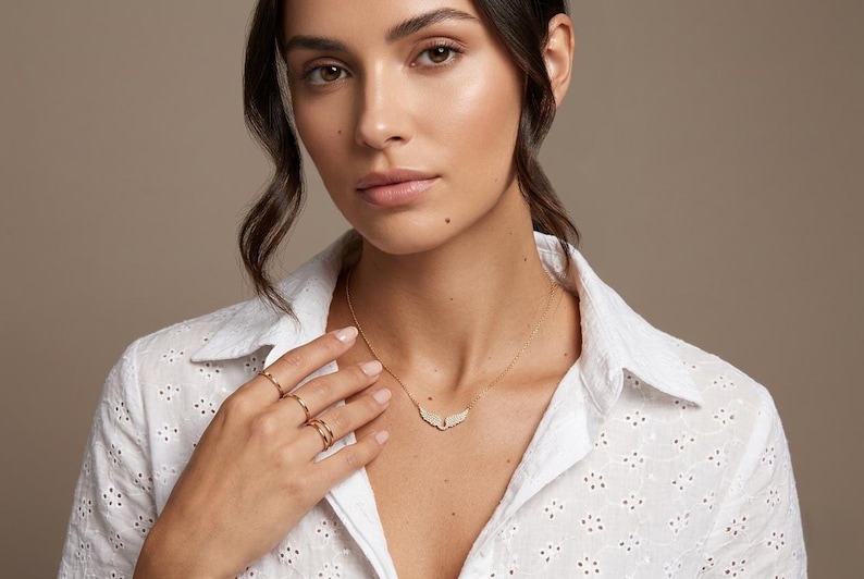 May include: A gold necklace with a wing-shaped pendant and several gold rings. The model is wearing a white eyelet shirt. The background is a solid, neutral color.