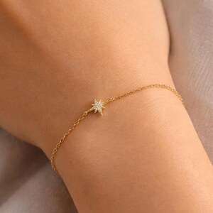 Könnte beinhalten: Ein zartes, goldfarbenes Armband mit einem Sternen-Charm mit kleinen, funkelnden Akzenten. Das Armband wird am Handgelenk getragen und zeigt seine feine Kette und das detaillierte Design des Sterns. Der Schmuck ist schlicht und elegant.