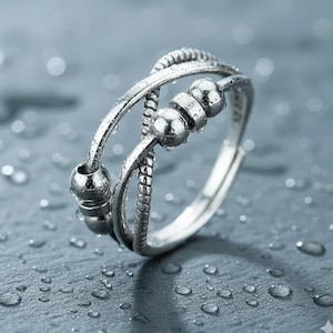 May include: A silver-toned ring with a textured design and small, movable beads. The ring features a braided rope-like pattern and a series of small, spherical beads that can be spun. The ring is shown against a wet, gray surface.