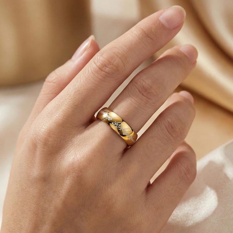 K&ouml;nnte beinhalten: Ein goldfarbener Ring mit einem eingravierten Muster. Der Ring wird an einem Finger getragen und zeigt sein detailliertes Design und seine polierte Oberfl&auml;che. Das Muster scheint eine Reihe stilisierter Bl&auml;tter oder Zweige zu sein. Der warme Farbton des Rings kontrastiert mit dem Hautton.