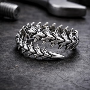 Puede incluir: Un anillo plateado con un diseño en forma de espina dorsal. El anillo presenta una serie de segmentos puntiagudos interconectados, creando una estética texturizada y atrevida. El diseño del anillo es detallado e intrincado, con un acabado pulido. Es una pieza de joyería única.