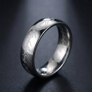 K&ouml;nnte beinhalten: Ein silberfarbener Ring mit eingravierter Schrift um das Band. Der Ring hat eine polierte, reflektierende Oberfl&auml;che, und die Schrift ist in einer stilisierten Schriftart gehalten. Der Ring ist wahrscheinlich aus Edelstahl oder einem &auml;hnlichen Metall gefertigt.
