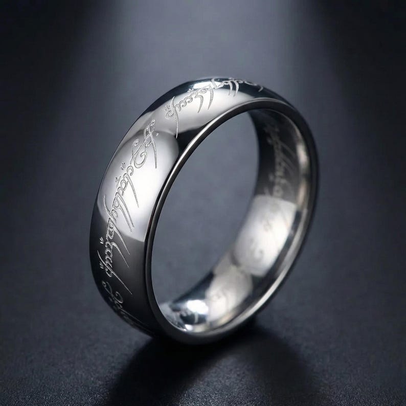 K&ouml;nnte beinhalten: Ein silberfarbener Ring mit eingravierter Schrift um das Band. Der Ring hat eine polierte, reflektierende Oberfl&auml;che, und die Schrift ist in einer stilisierten Schriftart gehalten. Der Ring ist wahrscheinlich aus Edelstahl oder einem &auml;hnlichen Metall gefertigt.