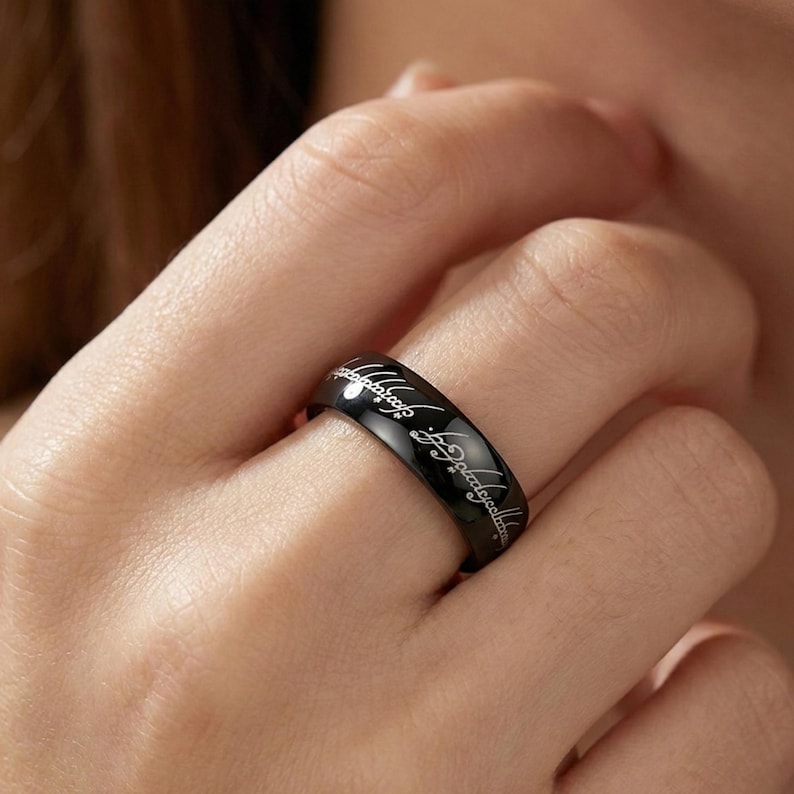 K&ouml;nnte beinhalten: Ein schwarzer Ring mit eingraviertem silbernem Text. Der Ring befindet sich an einem Finger. Der Text scheint in einer Schrift zu sein. Der Ring ist im Bandstil und hat ein gl&auml;nzendes Finish.