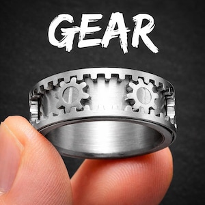 以下が含まれることがあります： 歯車デザインのシルバーリング。リングの上には白で「GEAR」の文字。暗い背景に、人の指でリングが持たれています。リングには、周囲に一連の歯車型の切り抜きがあります。