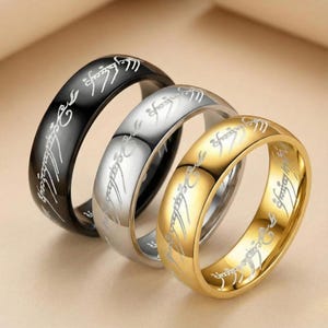 K&ouml;nnte beinhalten: Drei Ringe in Schwarz, Silber und Gold, jeweils mit eingravierter Schrift. Die Ringe werden schr&auml;g dargestellt, wodurch die detaillierte Inschrift um die B&auml;nder herum zur Geltung kommt. Der goldene Ring ist der hellste.