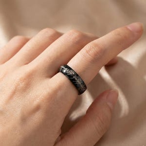 K&ouml;nnte beinhalten: Ein schwarzer Ring mit eingravierter Schrift. Der Ring wird an einem Finger getragen. Die Schrift ist in einer stilisierten Schriftart. Der Ring besteht aus einem gl&auml;nzenden, dunklen Material.