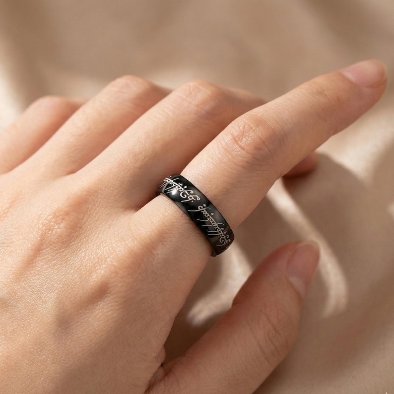 K&ouml;nnte beinhalten: Ein schwarzer Ring mit eingravierter Schrift. Der Ring wird an einem Finger getragen. Die Schrift ist in einer stilisierten Schriftart. Der Ring besteht aus einem gl&auml;nzenden, dunklen Material.