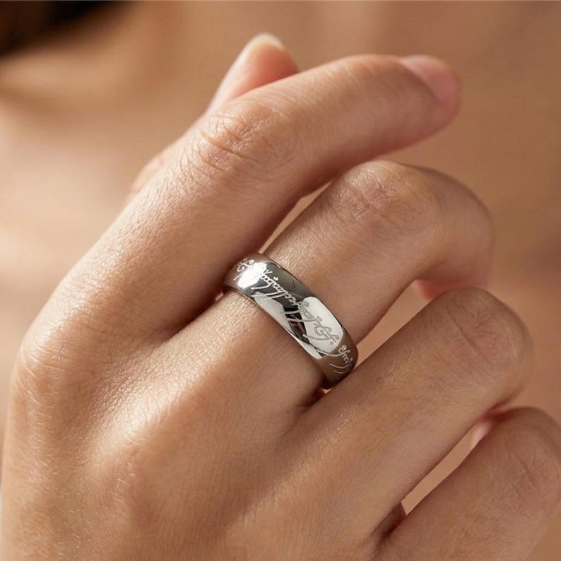 K&ouml;nnte beinhalten: Ein silberner Ring mit eingravierter Schrift. Der Ring wird an einem Finger getragen und zeigt seine polierte Oberfl&auml;che und detaillierte Inschrift. Das Design deutet auf ein Fantasy- oder literarisches Thema hin.