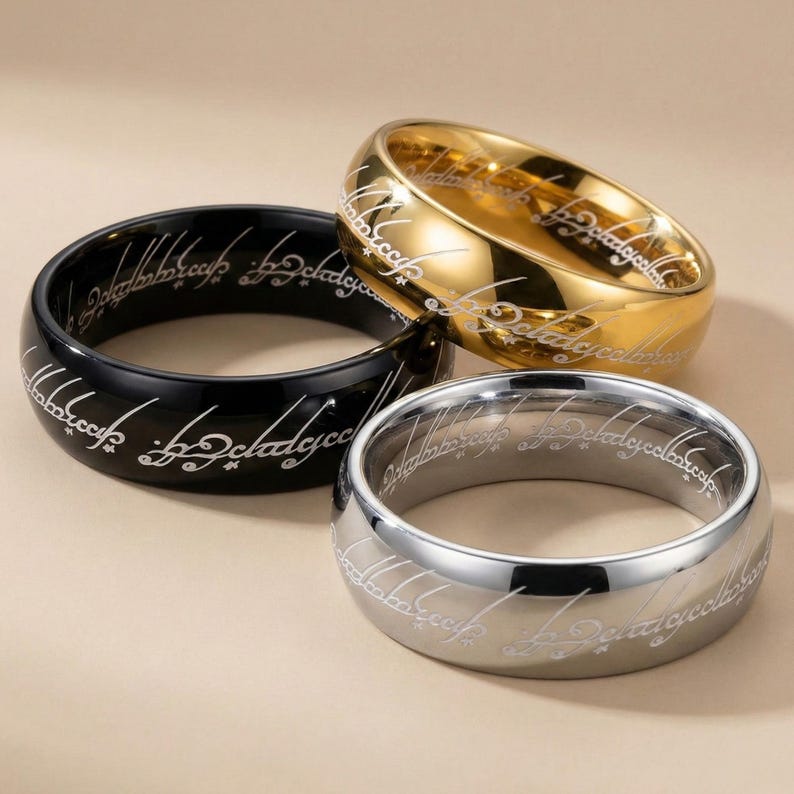 K&ouml;nnte beinhalten: Drei Ringe in Gold, Silber und Schwarz, jeweils mit eingravierter Schrift um das Band. Der goldene Ring hat eine gl&auml;nzende Oberfl&auml;che, der silberne Ring hat eine matte Oberfl&auml;che und der schwarze Ring hat eine gl&auml;nzende Oberfl&auml;che. Die Ringe sind wahrscheinlich von einem Fantasy-Thema inspiriert.