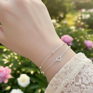 May include: Two delicate silver bracelets. The top bracelet features small beads, while the bottom one has a heart-shaped charm. The bracelets are worn on a wrist, with a blurred background of green foliage and pink flowers.