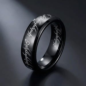 K&ouml;nnte beinhalten: Ein schwarzer Ring mit eingravierten wei&szlig;en Buchstaben. Der Ring hat eine polierte, reflektierende Oberfl&auml;che. Die Inschrift ist eine stilisierte Schrift. Der Ring ist ein Schmuckst&uuml;ck.