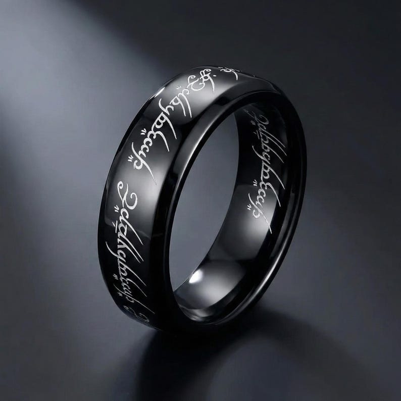 K&ouml;nnte beinhalten: Ein schwarzer Ring mit eingravierten wei&szlig;en Buchstaben. Der Ring hat eine polierte, reflektierende Oberfl&auml;che. Die Inschrift ist eine stilisierte Schrift. Der Ring ist ein Schmuckst&uuml;ck.