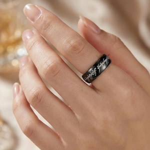 K&ouml;nnte beinhalten: Ein schwarzer Ring mit eingravierter Schrift. Der Ring wird an einem Finger getragen. Die Schrift ist in einer stilisierten Schriftart gehalten. Der Ring besteht aus dunklem Metall und hat eine glatte Oberfl&auml;che.