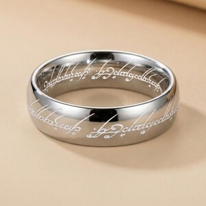 K&ouml;nnte beinhalten: Ein silberfarbener Ring mit wei&szlig;em graviertem Text um das Band. Der Ring hat eine polierte, reflektierende Oberfl&auml;che. Der Text ist in einer stilisierten Schrift. Der Ring ist ein Schmuckst&uuml;ck.