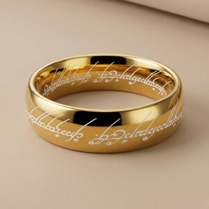 K&ouml;nnte beinhalten: Ein goldfarbener Ring mit wei&szlig;em, eingraviertem Text um das Band. Der Ring hat eine polierte Oberfl&auml;che und ein klassisches, abgerundetes Design. Der Text scheint in einer stilisierten Schrift zu sein. Der Ring ist wahrscheinlich ein Schmuckst&uuml;ck.
